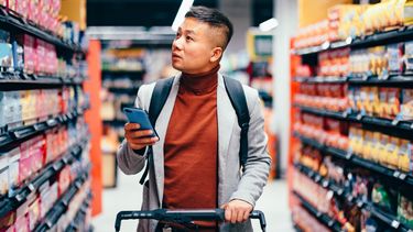 Asian male walking down the product aisle in the supermarket, looking at shelves and searching for groceries from the list on his mobile phone he is holding in his hand.