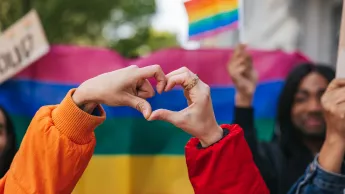 Genderqueere und nicht-binäre Freund*innen, die vor einer Regenbogenfahne eine Herzform bilden