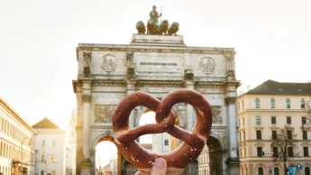 The girl is holding a delicious traditional German pretzel in the hand against the backdrop of the Victory Gate triumphal arch Siegestor in Munich.