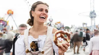 Junge Frau im Dirndl auf einem deutschen Volksfest. Sie hält eine Brezel in der Hand, im Hintergrund sieht man ein Riesenrad.