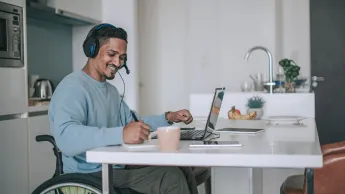 Adult Indian man in wheelchair working at home on laptop in his kitchen and has a video appointment.