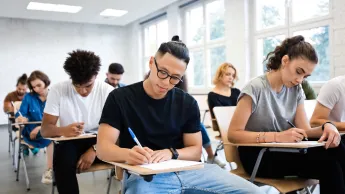 Young multiethnic students writing a test in the seminar room. In focus is a young Asian man with glasses.