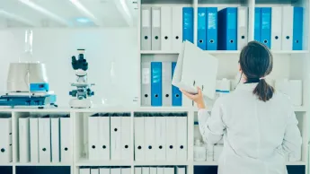 Woman in smock stands in front of a shelf with files, next to it is scientific laboratory equipment.