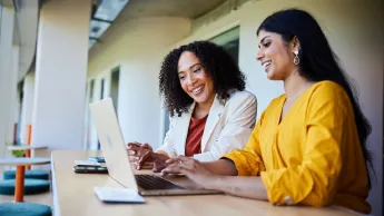 A Latin American mentor and an Asian mentee sit together at the table, smiling and talking.