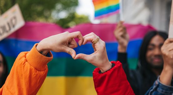 Genderqueere und nicht-binäre Freund*innen, die vor einer Regenbogenfahne eine Herzform bilden