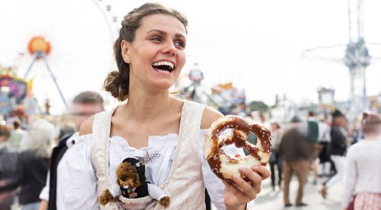 Junge Frau im Dirndl auf einem deutschen Volksfest. Sie hält eine Brezel in der Hand, im Hintergrund sieht man ein Riesenrad.